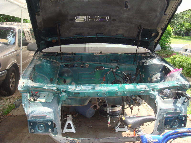 Green car engine bay with hood open, disassembled. SHO emblem visible on hood liner. Another vehicle in background