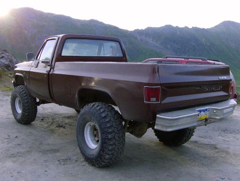 1982 Chevrolet C/K Pickup - A brown pickup truck with off-road tires is parked outdoors with mountains in the background.