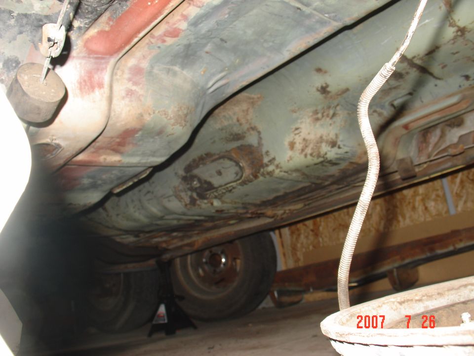 Underside of a vehicle showing rusty floorboards and some suspension parts. A wheel is visible on a jack stand