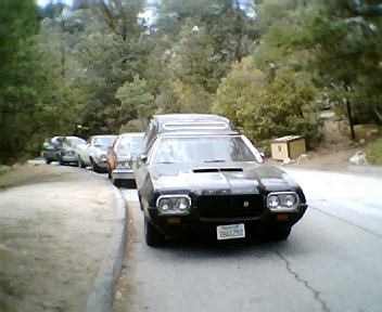 1972 Ford Ranchero - A line of vehicles, including a black 1972 Ford Ranchero, are parked on the side of a tree-lined road.