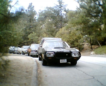 1972 Ford Ranchero - A black 1972 Ford Ranchero is seen from the front, driving on a road lined with trees. Other vehicles are visible in the background.