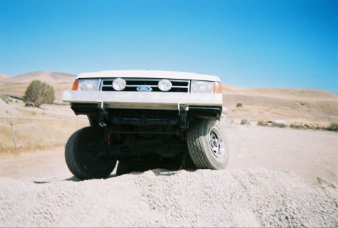 1991 Ford Bronco - A white 1991 Ford Bronco is seen from the front, navigating a gravelly incline under a bright blue sky.