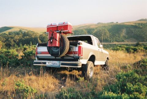 1991 Ford Bronco - Rear three-quarter view of a white full-size Ford Bronco parked in tall grass on a coastal hillside at golden hour, with the hardtop off an…