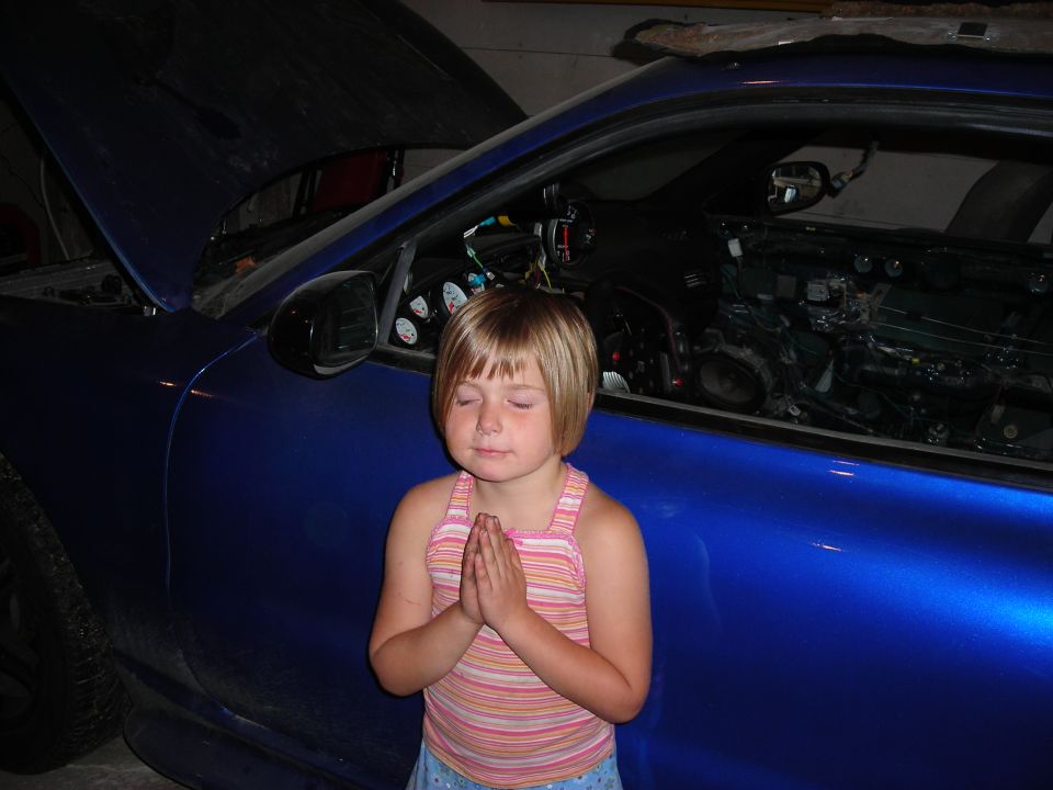 Young child posing with hands together in front of a blue car under repair