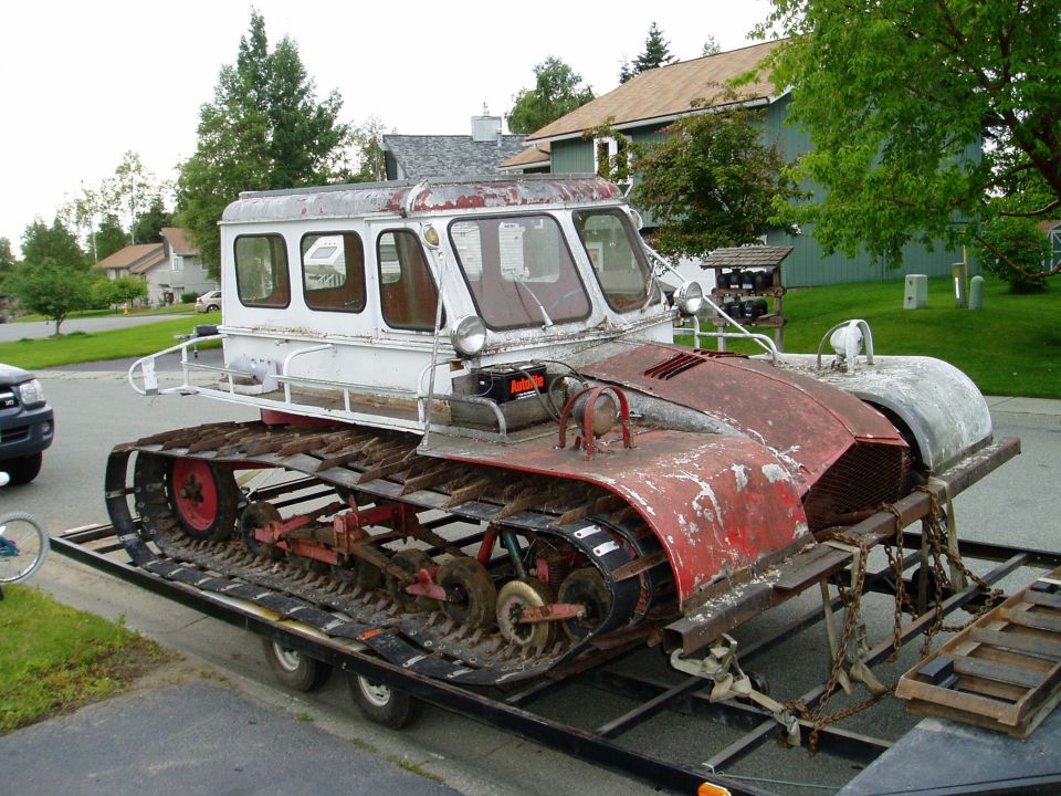 Snowcat vehicle with red and white body, tracks, and cabin, loaded on a trailer