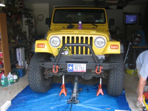 2001 Jeep Wrangler - Head-on front view of a yellow Jeep Wrangler TJ raised on orange jack stands over a blue tarp in a home garage, with a floor jack under the…