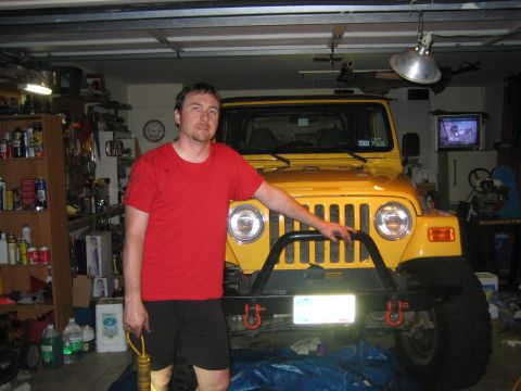 2001 Jeep Wrangler - A person in a red T-shirt stands beside the front of a yellow Jeep Wrangler TJ inside a cluttered home garage, resting a hand on the round…