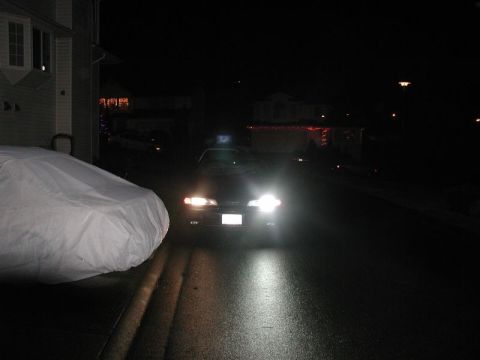 1990 Ford Thunderbird - A dark sedan is parked on a wet street at night, its headlights illuminating the road.
