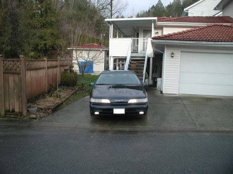 1990 Ford Thunderbird - A dark sedan is parked in a driveway, facing the camera with its headlights illuminated.