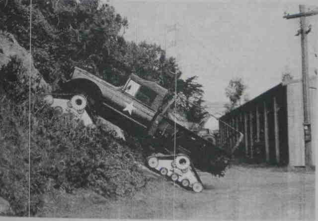 Truck with tank treads on a steep, wooded incline next to a building