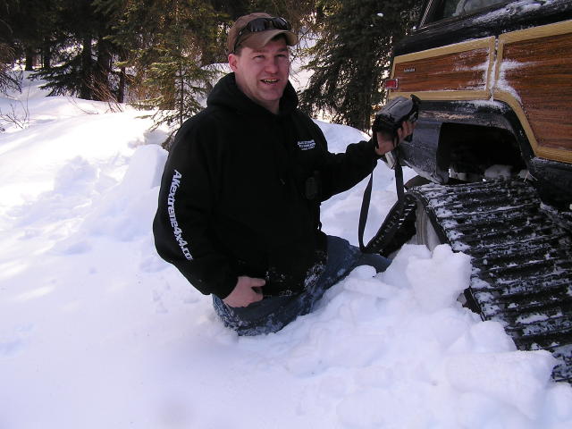 Guy in snow next to a vehicle with tracks and wood paneling. He's holding a camera