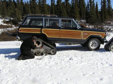 2000 Ford E350 - A person works on the snow track of a black Jeep Grand Wagoneer with wood paneling in a snowy, wooded area.
