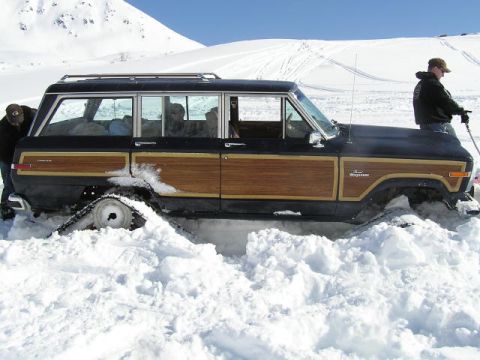 2000 Ford E350 - Wood-paneled Jeep Grand Wagoneer equipped with snow tracks is partially buried in a snowy landscape.