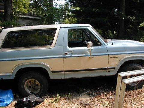 1986 Ford Bronco - This two-tone Ford Bronco is parked in a wooded area with some debris around it.