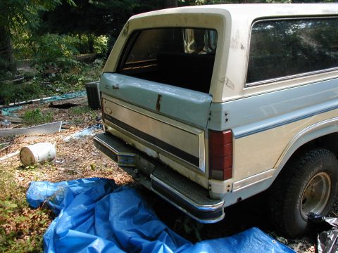 1986 Ford Bronco - The rear of a light blue and cream Ford Bronco is visible, parked outdoors with a blue tarp in front.