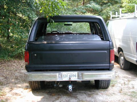 1986 Ford Bronco - The rear of a black Ford Bronco is visible, parked next to a white van in a wooded area.