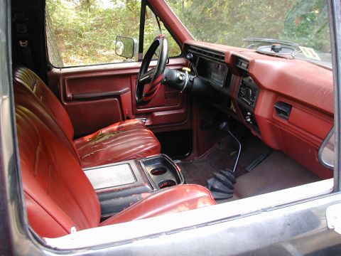 1986 Ford Bronco - The interior of a truck, red vinyl seats and a dashboard, is viewed from the passenger side.