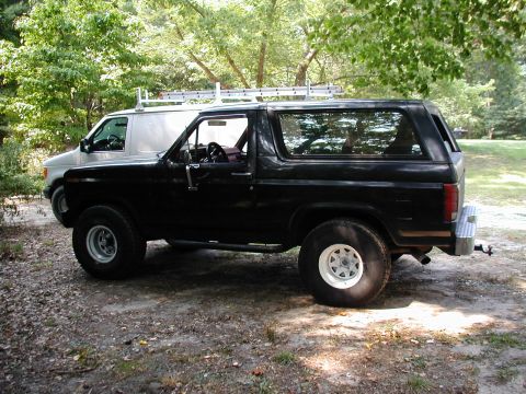 1986 Ford Bronco - A black 1986 Ford Bronco is parked outdoors next to a silver van.