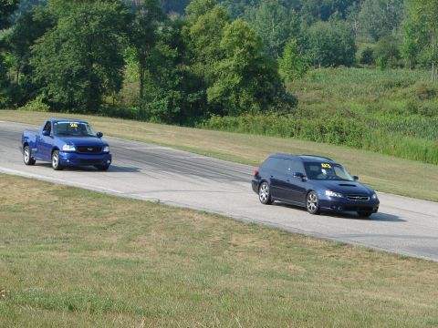 1993 Ford Taurus - Two vehicles, a blue pickup truck and a blue station wagon, are on a race track with grassy hills in the background.