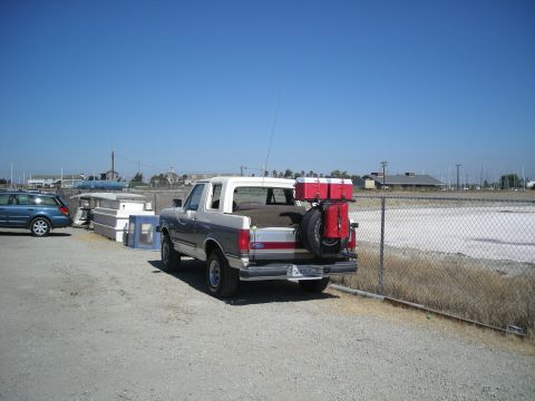 1991 Ford Bronco - Driver-side rear three-quarter view of a two-tone tan-over-gray full-size Ford Bronco with a white roof and red pinstripe, parked on gravel…