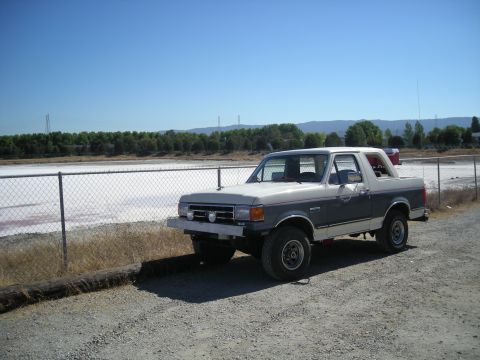 1991 Ford Bronco - A two-tone Ford Bronco is parked on a gravel lot with a body of water and trees in the background.