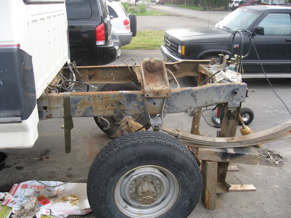Bare rusty rear frame of a pickup with bed removed, wheel and leaf spring on wooden supports in a driveway