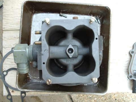 1978 Ford Bronco - A gray metal carburetor part, marked BI-27, rests in a metal tray with some screws and a gasket nearby.