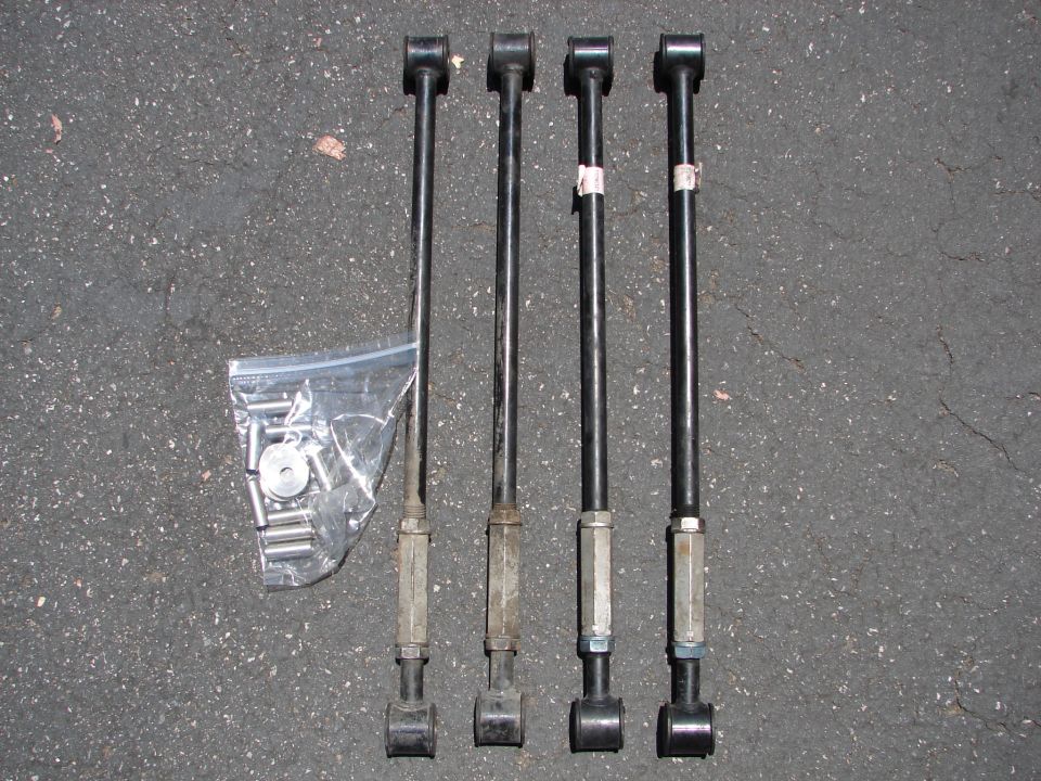 Four black suspension control arms and a bag of hardware laid out on asphalt
