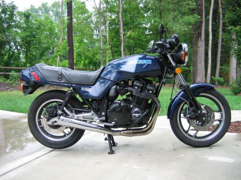 1985 Ford LTD Crown Victoria - Right-side profile of a dark blue Suzuki GS1100E standard motorcycle on its centerstand on a concrete driveway with green woods behind. It…