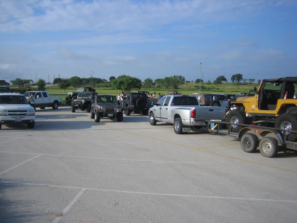 Several Jeeps and trucks parked in a lot, one Jeep is on a trailer being towed by a pickup