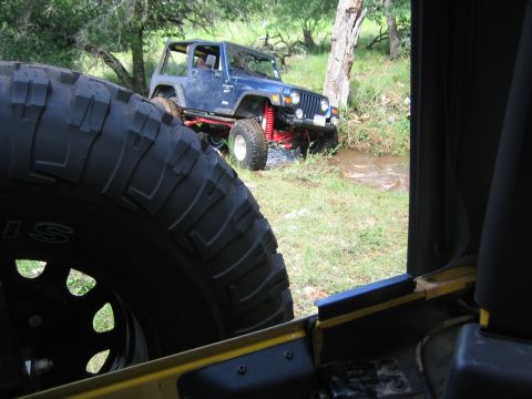 2001 Jeep Wrangler - A blue 2001 Jeep Wrangler navigates a stream with its red suspension visible, while another Jeep's tire is in the foreground.