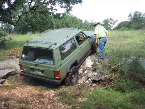 2001 Jeep Wrangler - A green Jeep Cherokee XJ is shown off-roading over rocks and dirt, with a person observing.
