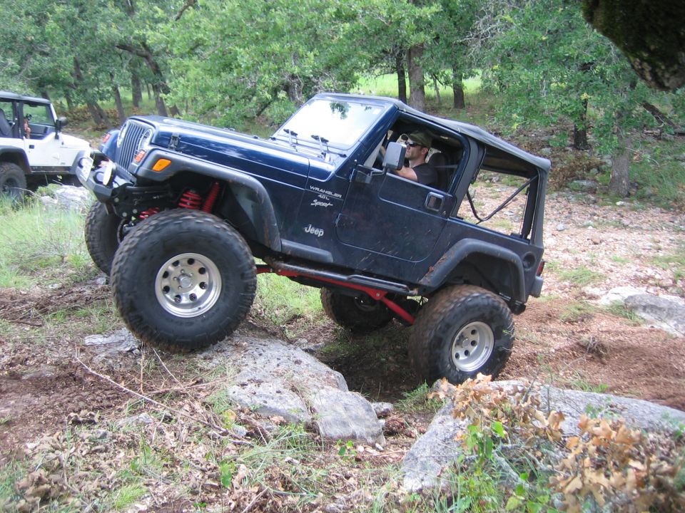 Blue Jeep Wrangler with lifted suspension driving over rocks on a trail. Another white Jeep is visible in the background
