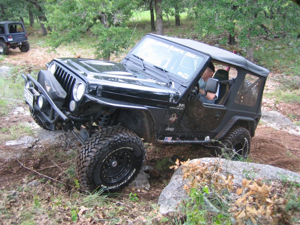 Black Jeep Wrangler on rocky terrain, front-left tire angled up, driver visible