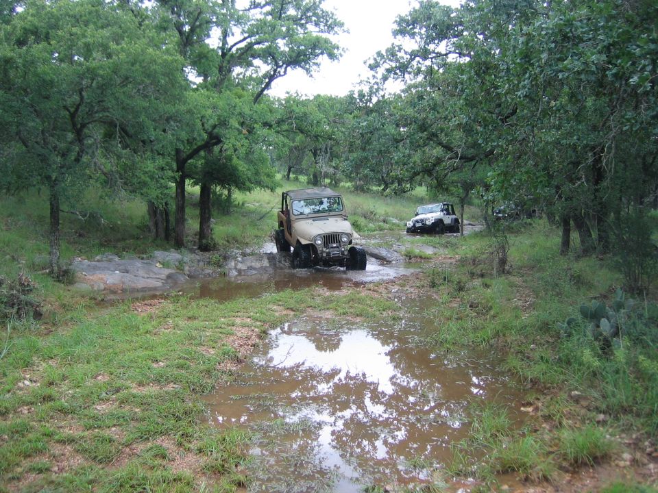 Tan Jeep Wrangler driving through a muddy puddle, with a white Jeep and another vehicle behind it in a wooded area