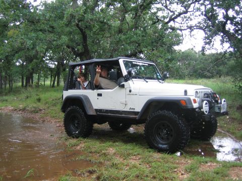 2001 Jeep Wrangler - A white Jeep Wrangler navigates a muddy path surrounded by trees, with two occupants enjoying the ride.