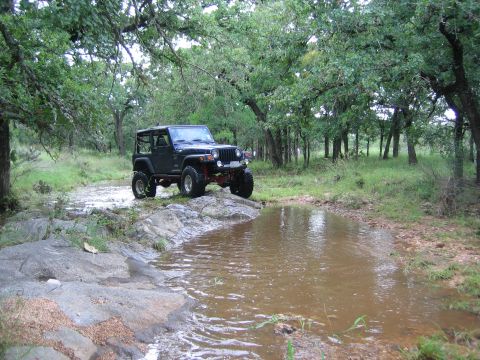 2001 Jeep Wrangler - A black Jeep Wrangler navigates a muddy path through a wooded area with a small body of water.