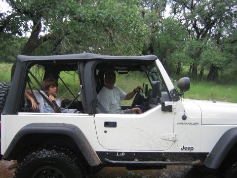 2001 Jeep Wrangler - Two people are inside a white Jeep Wrangler as it drives through muddy water with trees in the background.
