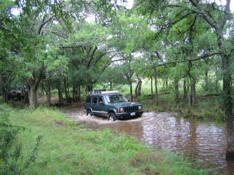 2001 Jeep Wrangler - A Jeep Cherokee XJ navigates a water crossing surrounded by lush green trees and foliage.