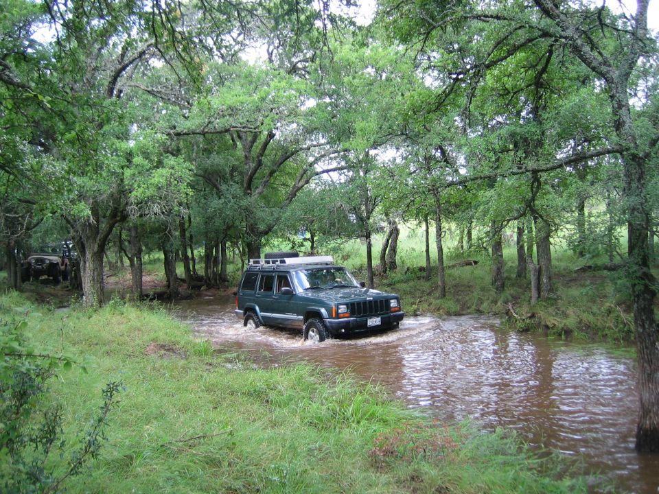 Green Jeep Cherokee XJ driving through a shallow stream in a wooded area. Another vehicle is visible in the background