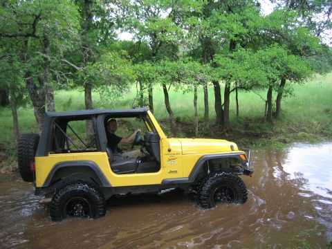 2001 Jeep Wrangler - A yellow 2001 Jeep Wrangler navigates through a shallow body of water surrounded by trees.