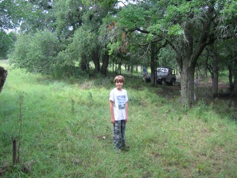 2001 Jeep Wrangler - A young boy stands in a grassy clearing surrounded by trees, with two off-road vehicles parked in the background.