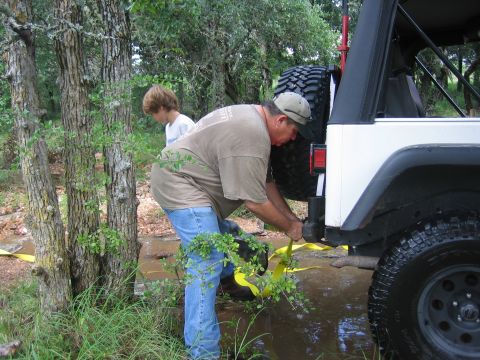 2001 Jeep Wrangler - A man attaches a yellow tow strap to the hitch of a white Jeep Wrangler, with a young boy standing nearby in a wooded area.