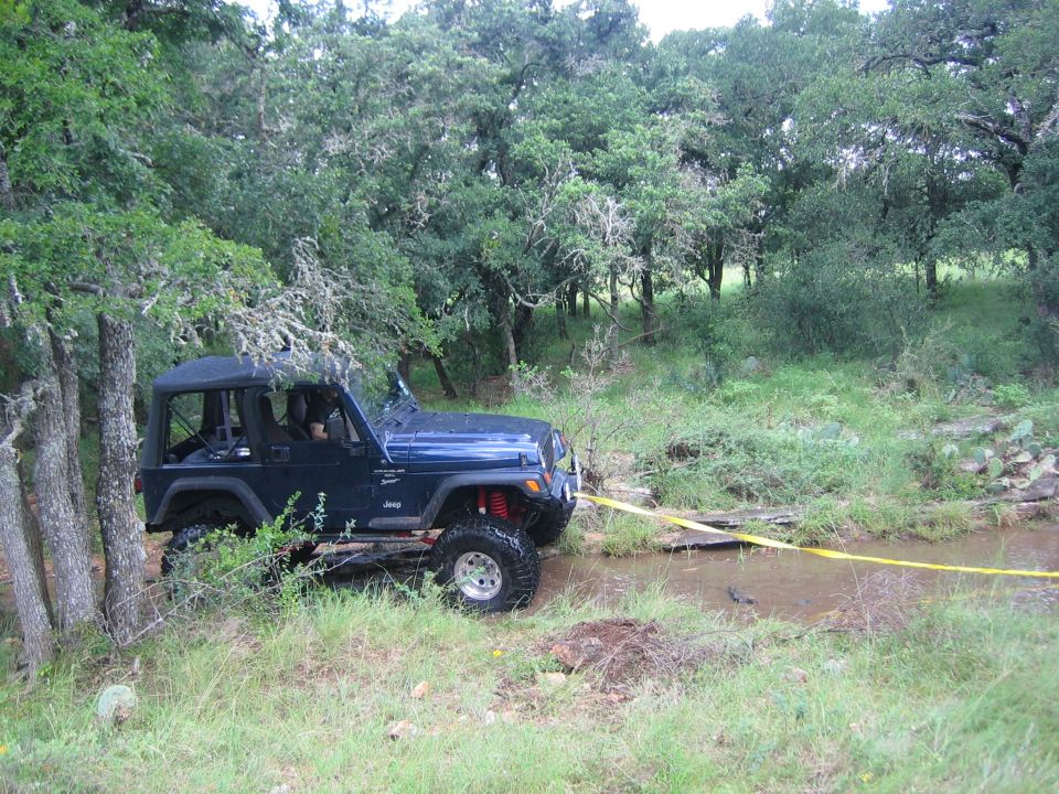 Blue Jeep Wrangler with yellow tow strap crossing a shallow stream in a wooded area