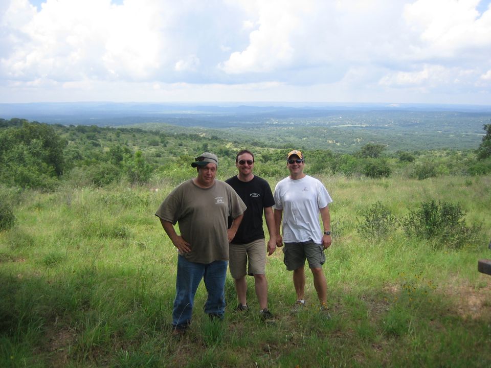 Three men stand in a grassy field overlooking a vast, tree-covered landscape under a cloudy sky