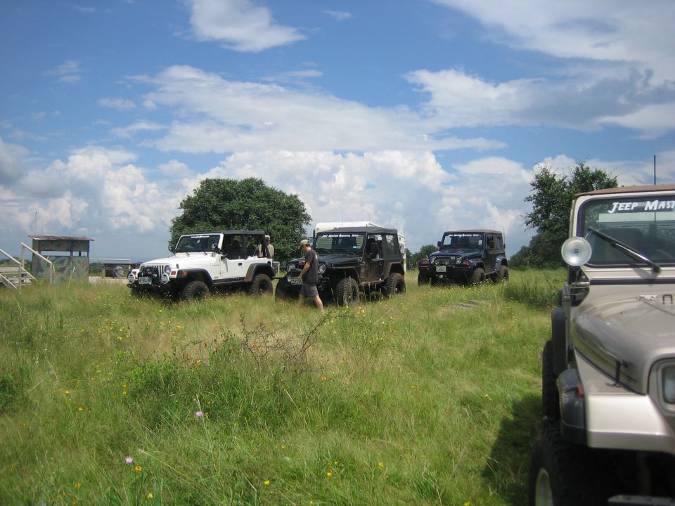 Four Jeep Wranglers parked in a grassy field under a cloudy sky. One white, one black, one dark blue, and one tan