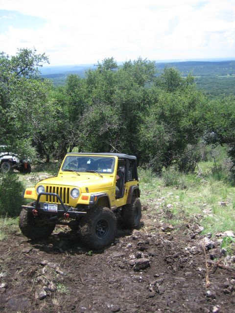 2001 Jeep Wrangler - A yellow 2001 Jeep Wrangler navigates a muddy off-road trail with a scenic overlook in the distance.