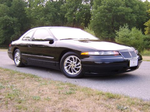 1996 Mercury Grand Marquis - A black coupe with distinctive chrome wheels is parked on a paved road with a wooded area behind it.