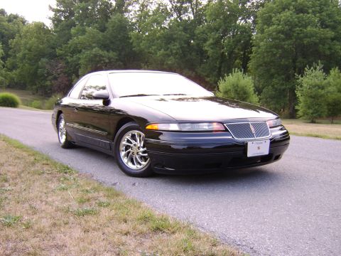 1996 Mercury Grand Marquis - This black coupe with chrome wheels is parked on the side of a road with trees in the background.