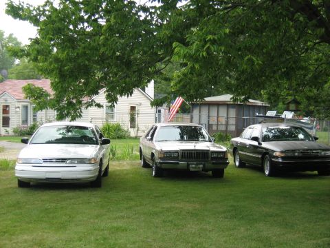 1988 Mercury Grand Marquis - Three sedans are parked on a lawn in front of houses, with one being a 1988 Mercury Grand Marquis.
